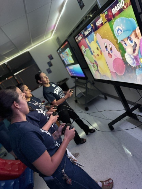 Three people play video games on a large screen in a room with multiple screens.