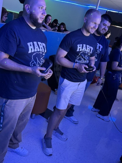 Individuals stand in a gaming area. Two men play, each holding a controller, with one wearing a blue shirt and khaki pants. The other wears white shorts.