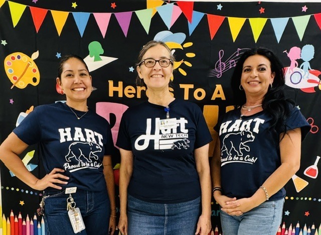 Three women wearing navy blue shirts with the word Hart and standing in front of a colorful banner.