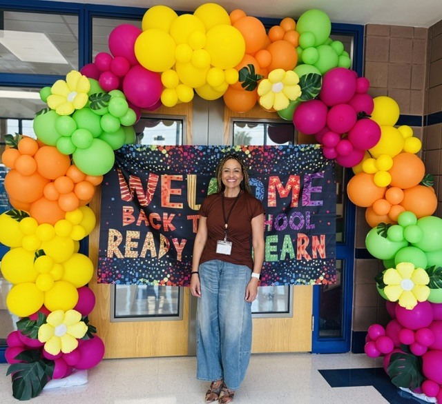 Person stands in front of colorful balloon arch with "Welcome Back To School Ready To Learn" sign.
