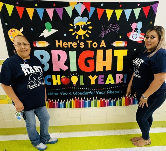 Two women stand smiling in front of a colorful banner with text that says "Here's to a bright school year!"