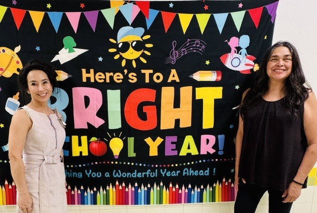 Two women stand beside a colorful banner that says, "Here's To A Right School Year!" in front of a classroom.