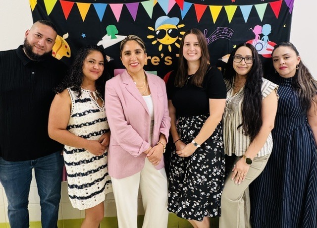 A group of six people, including two women wearing glasses, are standing together in front of a colorful banner.