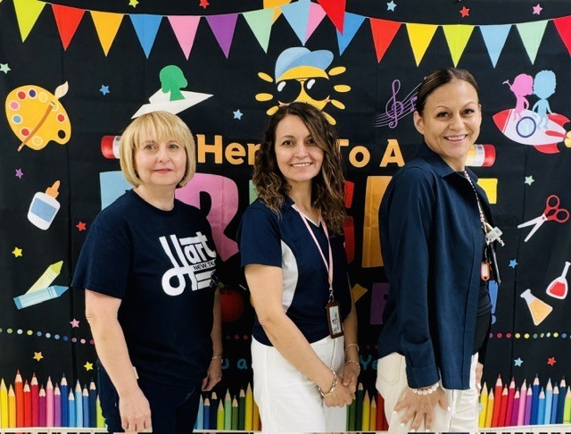 Three women stand in front of a colorful backdrop with a banner saying "Here to a B."