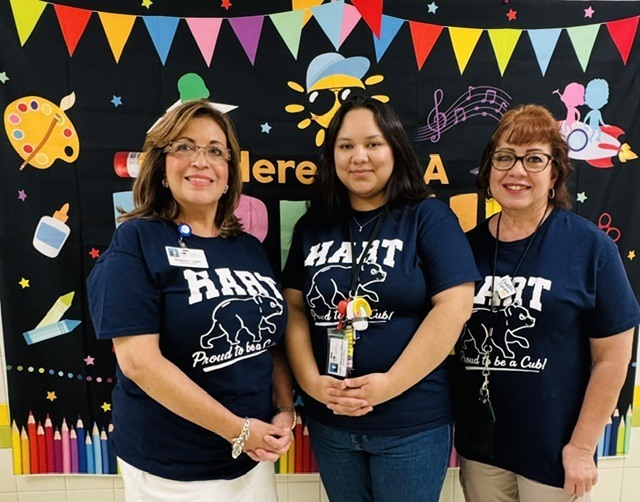 Three women in navy blue shirts, standing side by side in front of a vibrant, colorful classroom bulletin board.