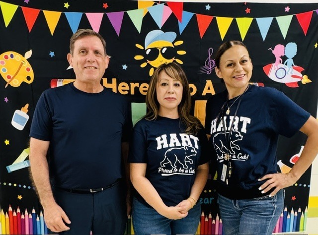 Three people stand in front of a colorful banner with a drawing of a bee and a sun.