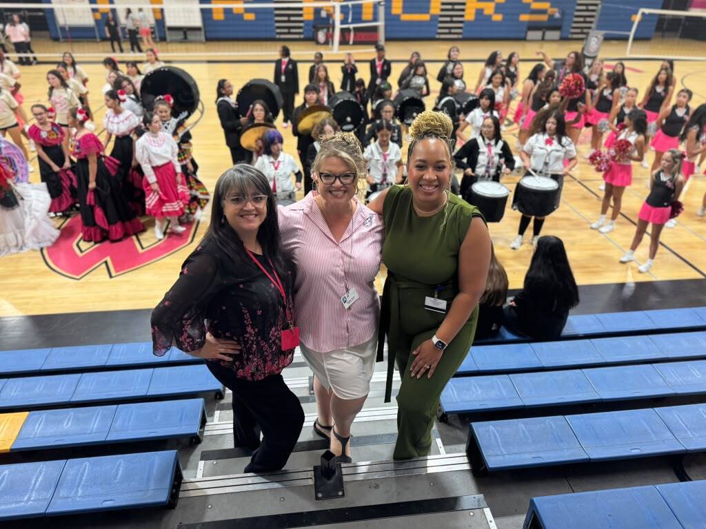 Three women stand in a gym. One wears glasses. They face the camera with a crowd behind them.