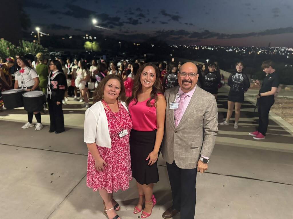Three individuals pose for a photo, smiling. They stand on a concrete surface with a group behind them.