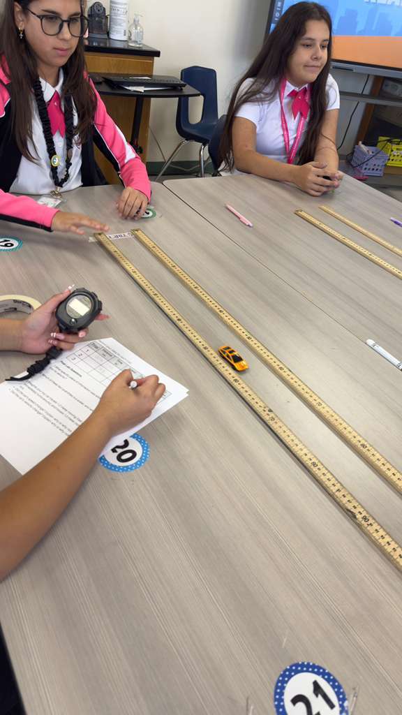 A group of students sit at a table. They have toy cars, a stopwatch, and measuring tapes.