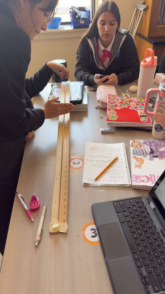 Two students work together at a table with various school supplies. One holds a ruler. The table includes a laptop, a notebook, and a cup.