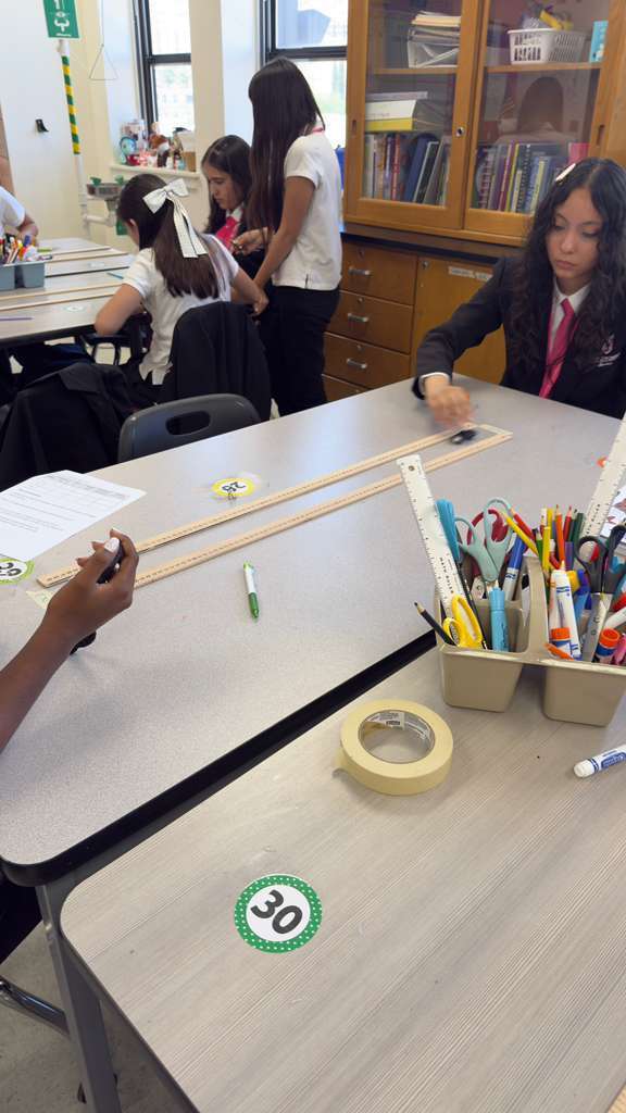 Five students, some seated and others standing, work on a project at desks. A ruler, tape, markers, and papers are on the tables.