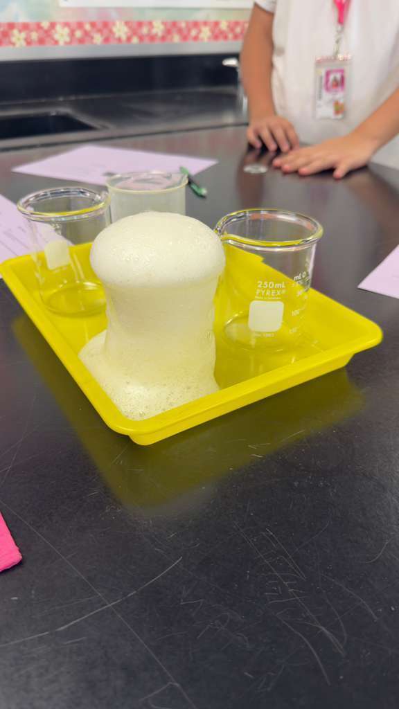 A science experiment involving a foamy substance sits on a yellow tray, with a person in a white shirt standing nearby.