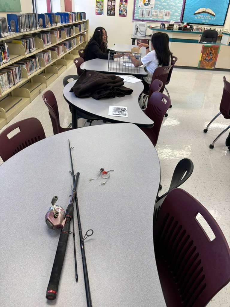 Two individuals seated at a table in a library with fishing rods on the table. Shelves with books in the background.