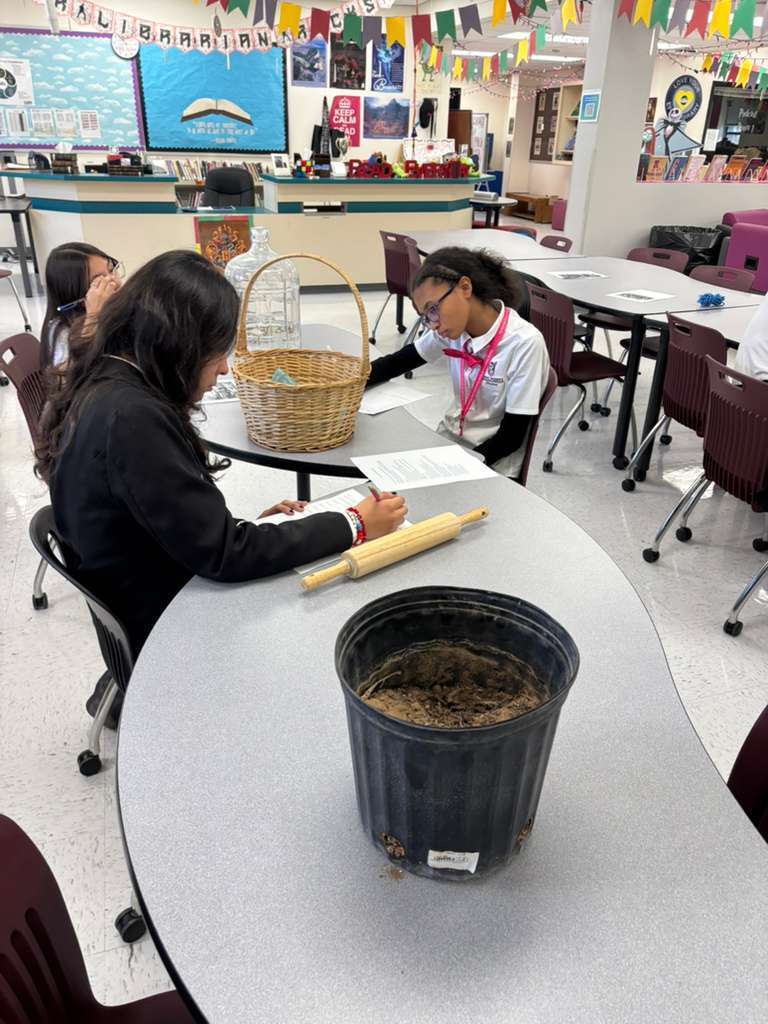 Students in a classroom sit at a table with a basket, a bucket, and a rolling pin.