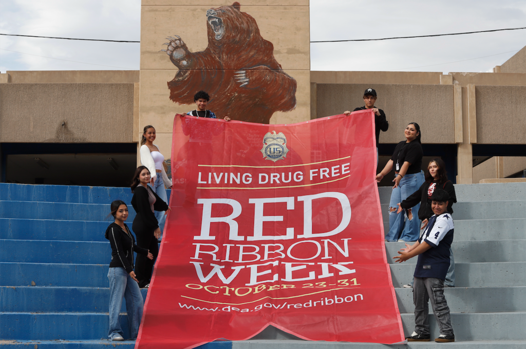 Bowie High School students smile for a photo with giant red banner that says Living Drug Free Red Ribbon Week October 23-31 www.dea.gov/redribbon