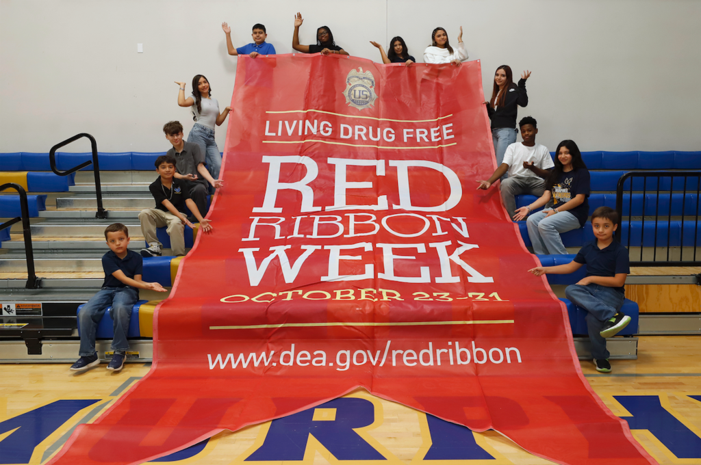 Murphree PK-8 School students smile for a photo with giant red banner that says Living Drug Free Red Ribbon Week October 23-31 www.dea.gov/redribbon