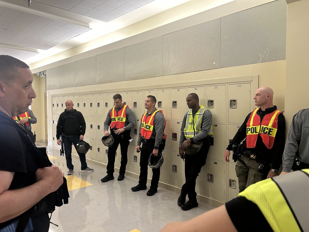 A group of police officers in safety vests labeled “POLICE” stand in a school hallway lined with lockers. Some hold helmets while others rest against the lockers, appearing to be in discussion or awaiting instructions. The setting is indoors with bright lighting and a tiled floor.