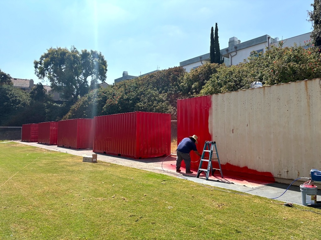 Person wearing a hat paints a large outdoor storage container bright red using a sprayer, with several other red-painted containers lined up in the background along a grassy field.