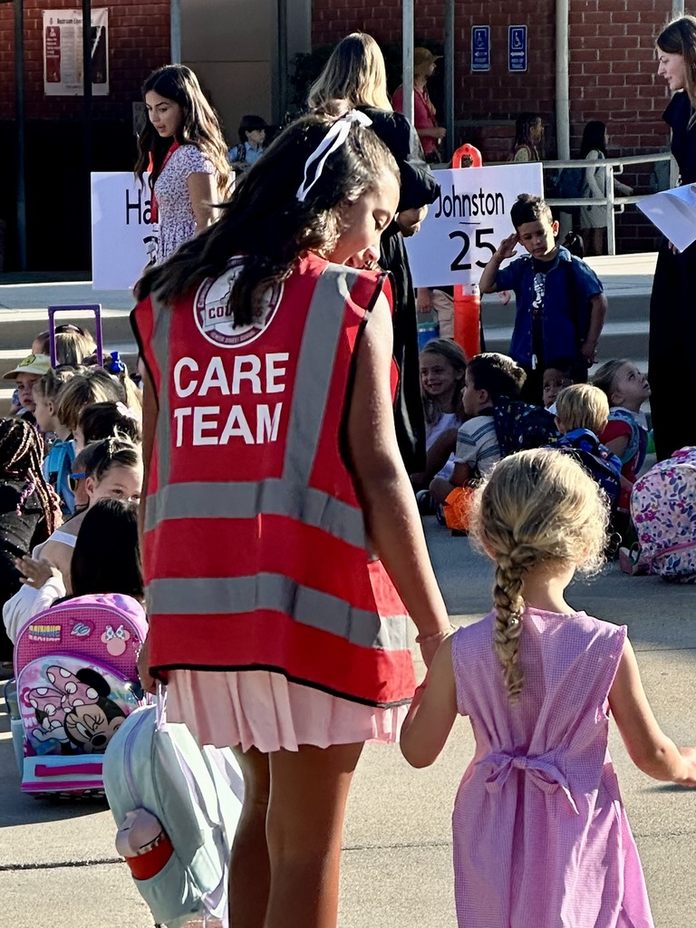 A student in a red vest labeled “CARE TEAM” holds hands with a younger child in a pink dress while walking through a group of seated students outside