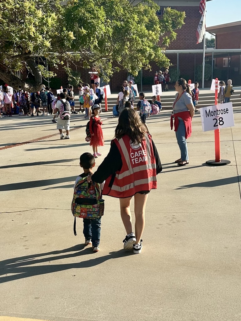 A student wearing a red vest labeled “CARE TEAM” walks with a younger child carrying a Mario-themed backpack. The two are heading toward a group of students and teachers gathered outside