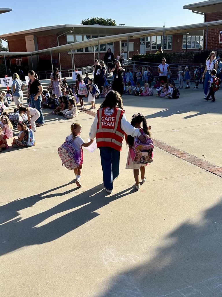 A student wearing a red vest labeled “CARE TEAM” walks hand in hand with two younger students carrying backpacks. They are surrounded by groups of children, parents, and staff gathered outside on the school grounds.
