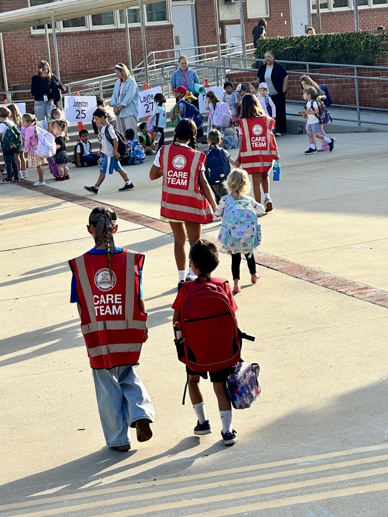 Two students wearing red vests labeled “CARE TEAM” walk alongside younger children carrying backpacks toward a group of classmates and teachers outside.