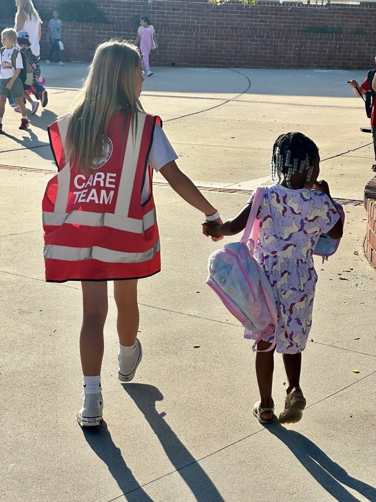 An older student wearing a red safety vest labeled “CARE TEAM” holds hands with a younger student carrying a unicorn backpack, walking together on a school playground. Other children and adults are visible in the background.