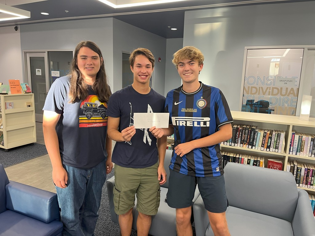 Three high school students stand together in a library, smiling and holding an envelope.