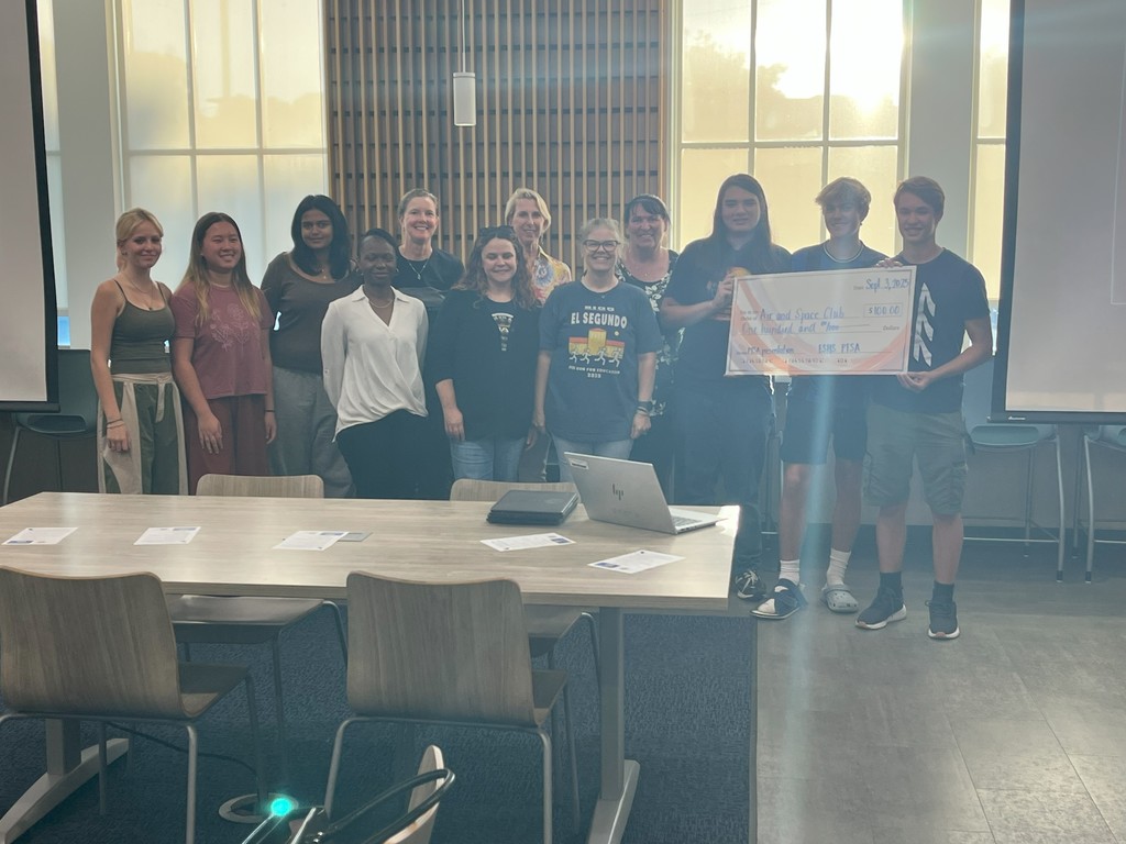 A group of students and adults pose together in a library, with three students holding a large ceremonial check made out to the Air and Space Club.