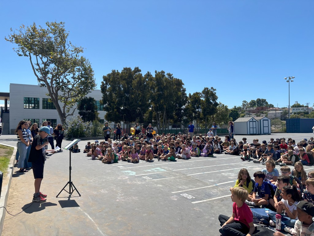 An elementary student wearing a blue hat reads from a podium with a microphone during an outdoor school assembly. Behind him, rows of students sit on the blacktop with staff members standing nearby.
