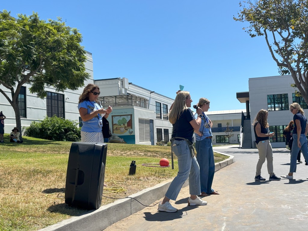 A staff member speaks into a microphone while standing outside near a grassy hill at a school. Another staff member nearby holds notes, and additional staff are standing in the background under a clear blue sky.