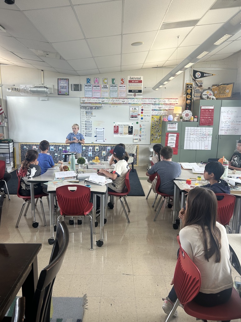 Elementary classroom with students seated at red chairs around group tables, facing the front of the room. A student stands at the whiteboard presenting while classmates listen. The whiteboard is covered with charts, posters, and classroom rules, and Pittsburgh Steelers decorations are visible on the side wall.