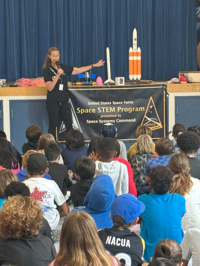 Presenter with a microphone stands on stage in front of a banner that reads “United States Space Force Space STEM Program presented by Space Systems Command.” She gestures toward rocket models while students watch from the floor.