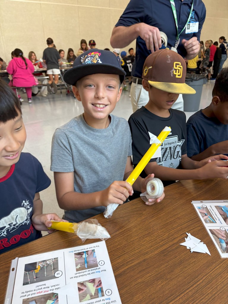 Smiling student wearing a baseball cap holds a yellow paper rocket at a table covered with paper instructions and supplies. Other students sit beside him working on their projects