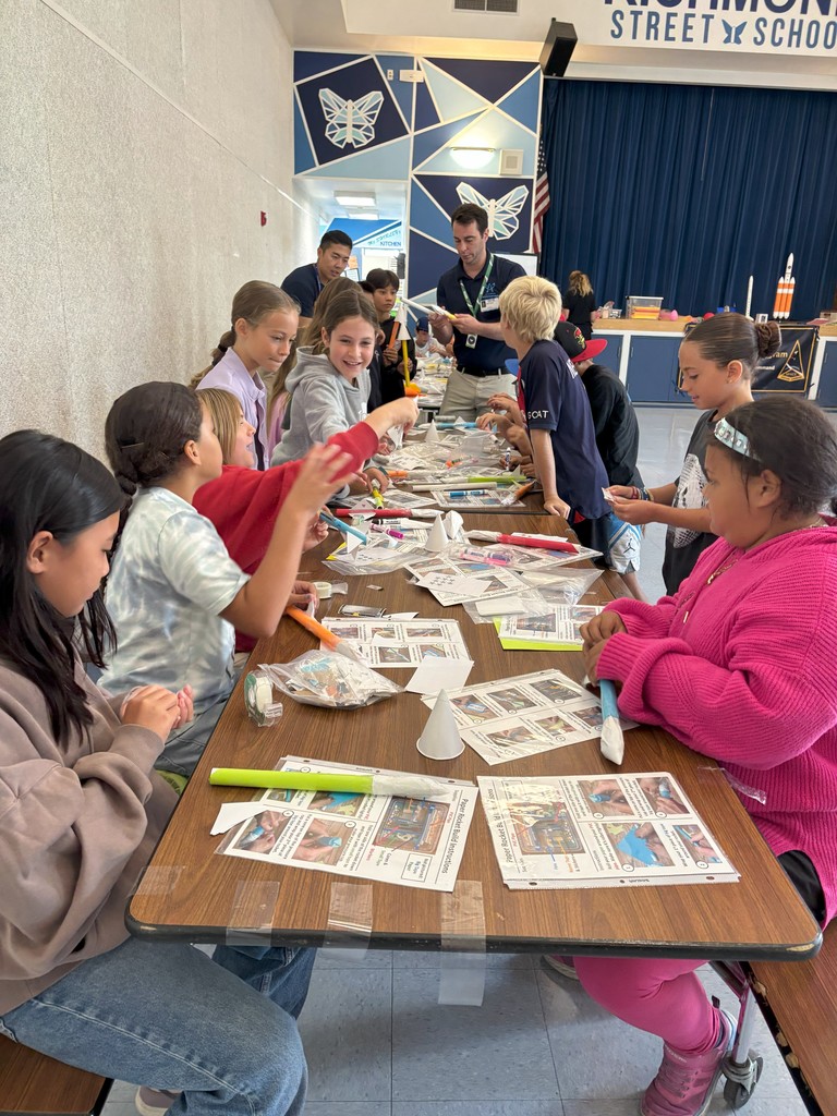 Group of students sit around a long table assembling paper rockets with tape and scissors. Instruction sheets and supplies are spread across the table while instructors walk around assisting.