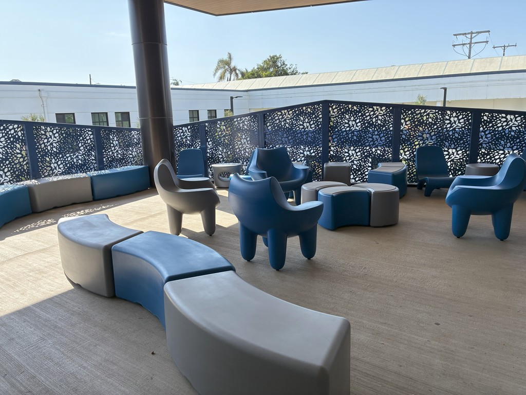 Outdoor seating area with modern blue and gray chairs and curved benches arranged on a covered patio. Decorative blue metal panels with floral cutouts line the railing.
