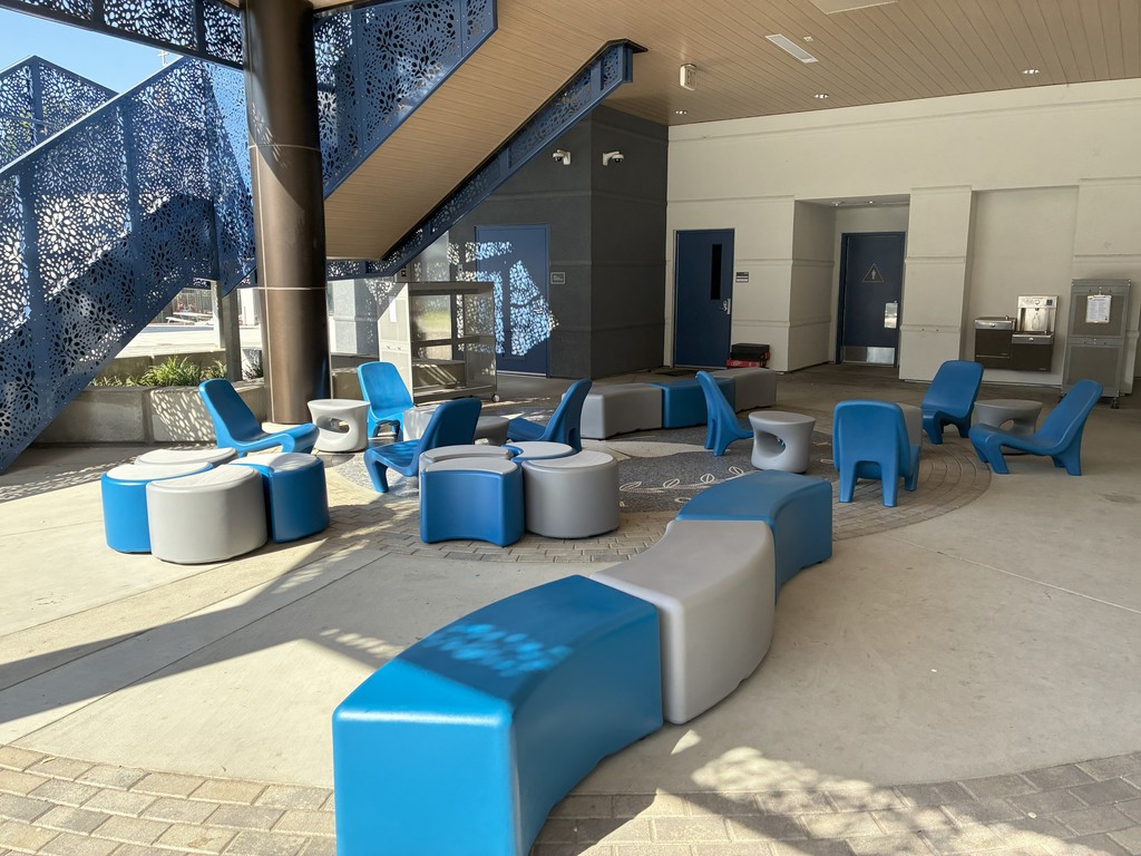 Shaded outdoor space with blue and gray lounge chairs, curved benches, and round stools arranged under a staircase. The area features decorative metal panels and access to building entrances.