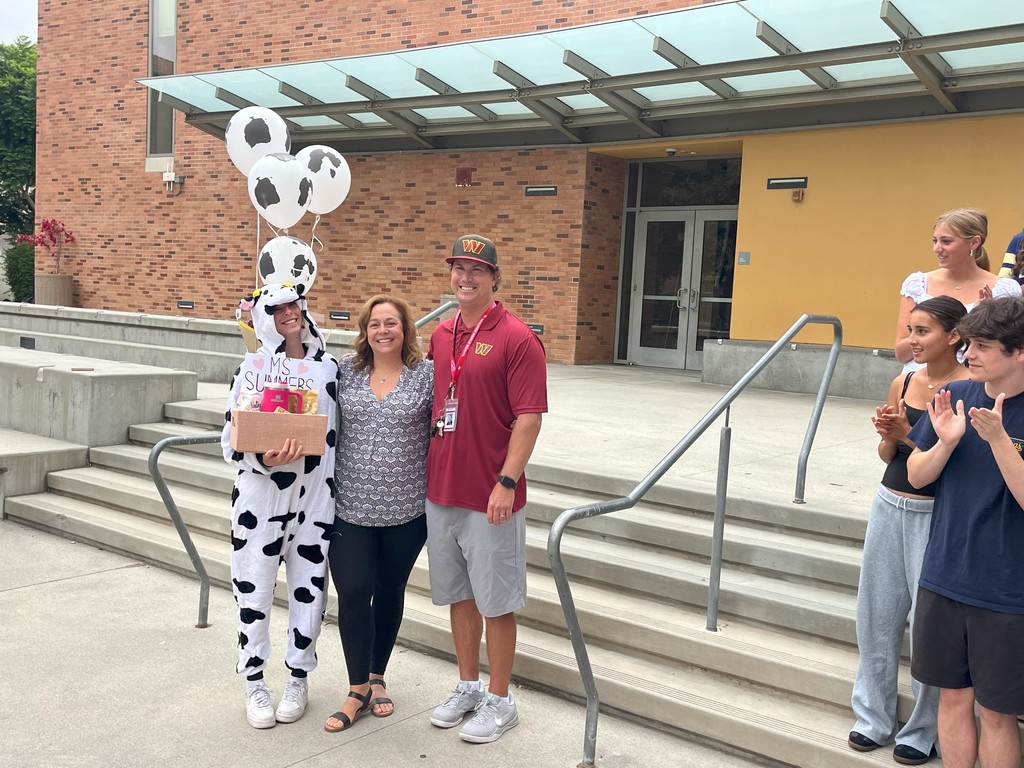 A student dressed in a cow costume holds balloons and a gift basket labeled “Ms. Summers,” standing with two adults smiling for a photo
