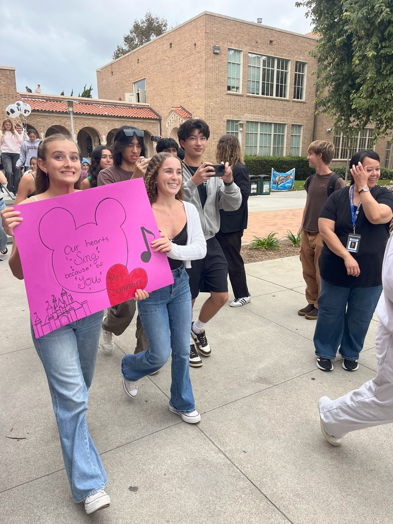 Students walk holding a large pink sign reading “Our hearts sing because of you Mrs. Summers,” while others record with phones and smile.