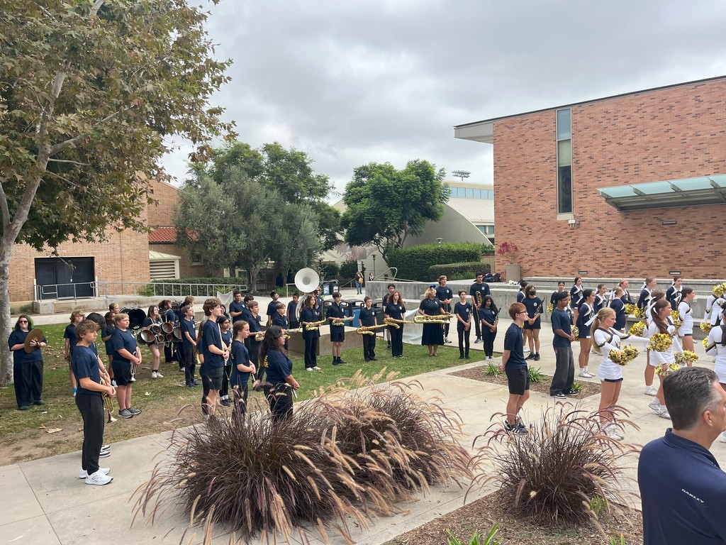 A marching band and cheerleaders perform in an outdoor courtyard, facing the crowd gathered on the steps.