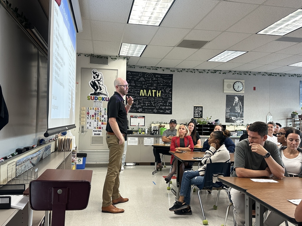 A teacher presents at the front of a classroom while parents sit at tables listening