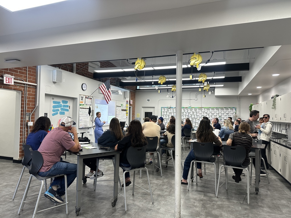 Adults sit at tall tables in a classroom-style lab while a presenter at the front points to a projected slide.