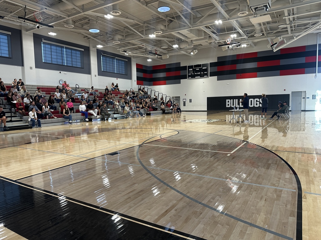 Staff members sit on bleachers in a school gym listening to a speaker near the court.
