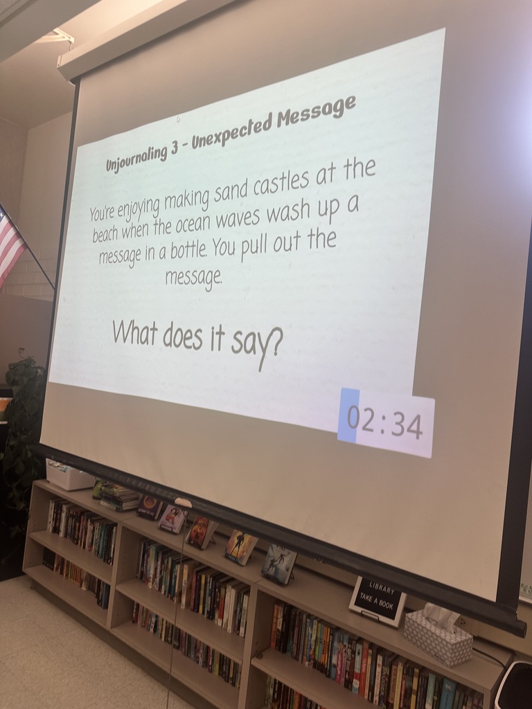 A classroom projector screen displays a writing prompt titled “Unjournaling 3 – Unexpected Message.” The text reads: “You’re enjoying making sand castles at the beach when the ocean waves wash up a message in a bottle. You pull out the message. What does it say?” A digital timer in the bottom right shows 2 minutes and 34 seconds.