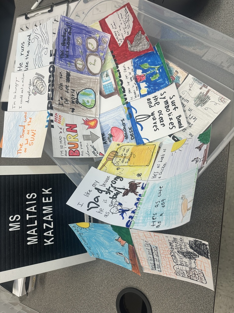A collection of colorful, hand-drawn student cards spread out in a clear bin. Each card features creative writing or illustrations, with phrases like “He runs as fast as the wind,” “Surf boards symbolize the oceans and waves,” and “I like my Dad because he is strong.” A black letter board next to the bin reads “Ms Maltais Kazamek.”