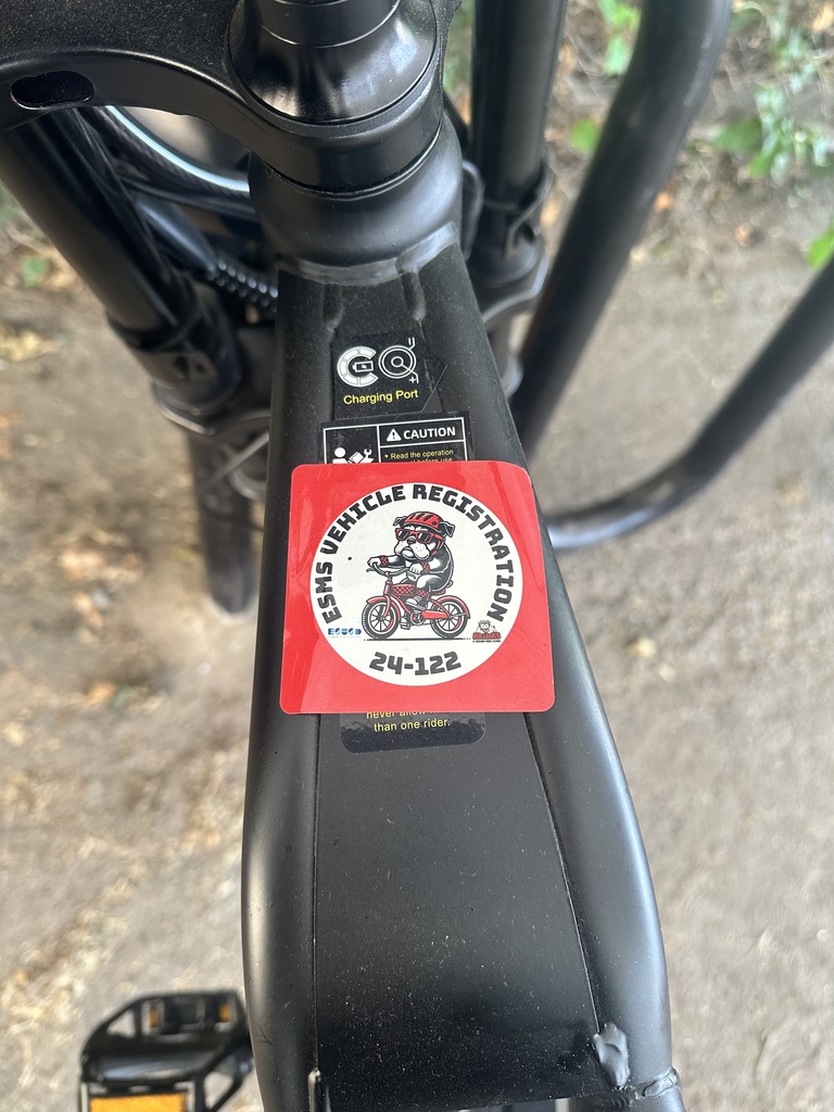 Close-up of a black electric bike frame showing a red sticker labeled “ESMS Vehicle Registration 24-122.” The sticker features a cartoon bulldog mascot riding a red bicycle.