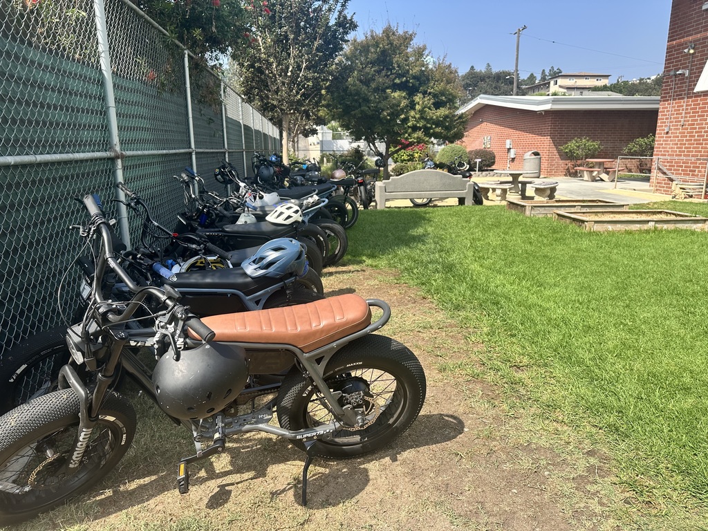 Row of electric bikes and helmets parked along a chain-link fence beside a school building.