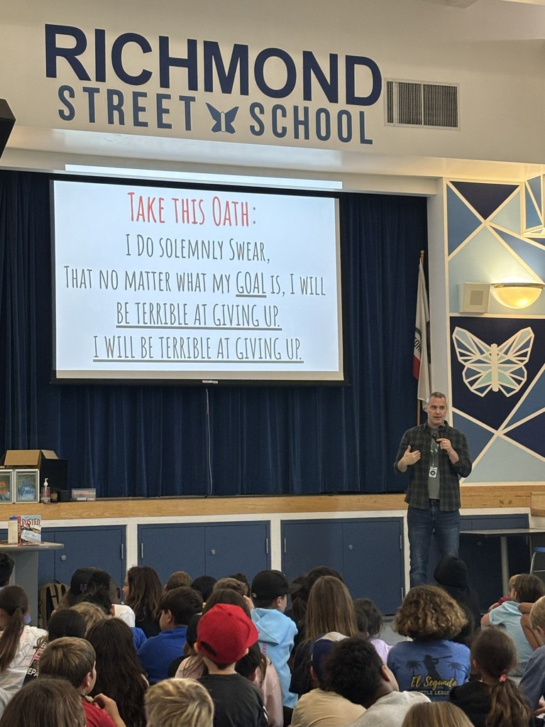 The presenter stands on stage speaking into a microphone as students sit facing him. The screen behind him displays a pledge that begins with “Take this Oath: I do solemnly swear…” beneath the Richmond Street School sign.