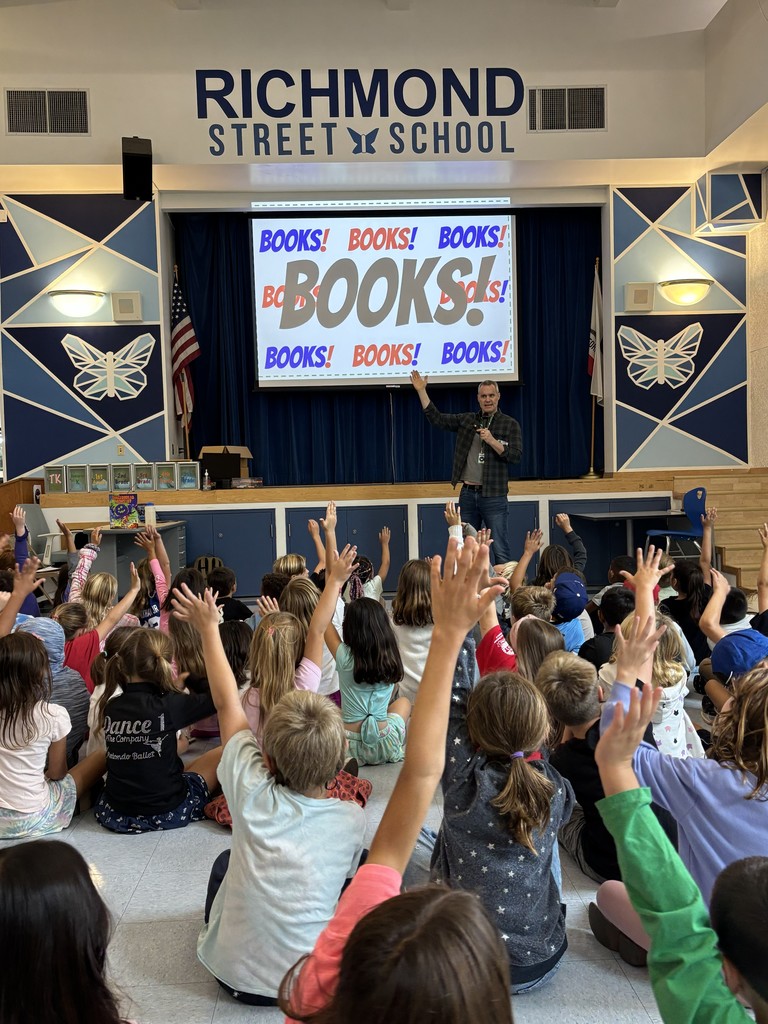 Students seated on the floor enthusiastically raise their hands toward the stage as the presenter speaks. The screen behind him reads “BOOKS!” in large letters repeatedly, and the Richmond Street School sign is visible above the stage.