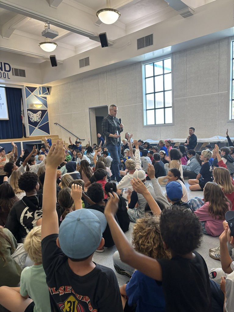 A wide view of the auditorium shows the presenter walking among seated students, many with their hands raised to participate.